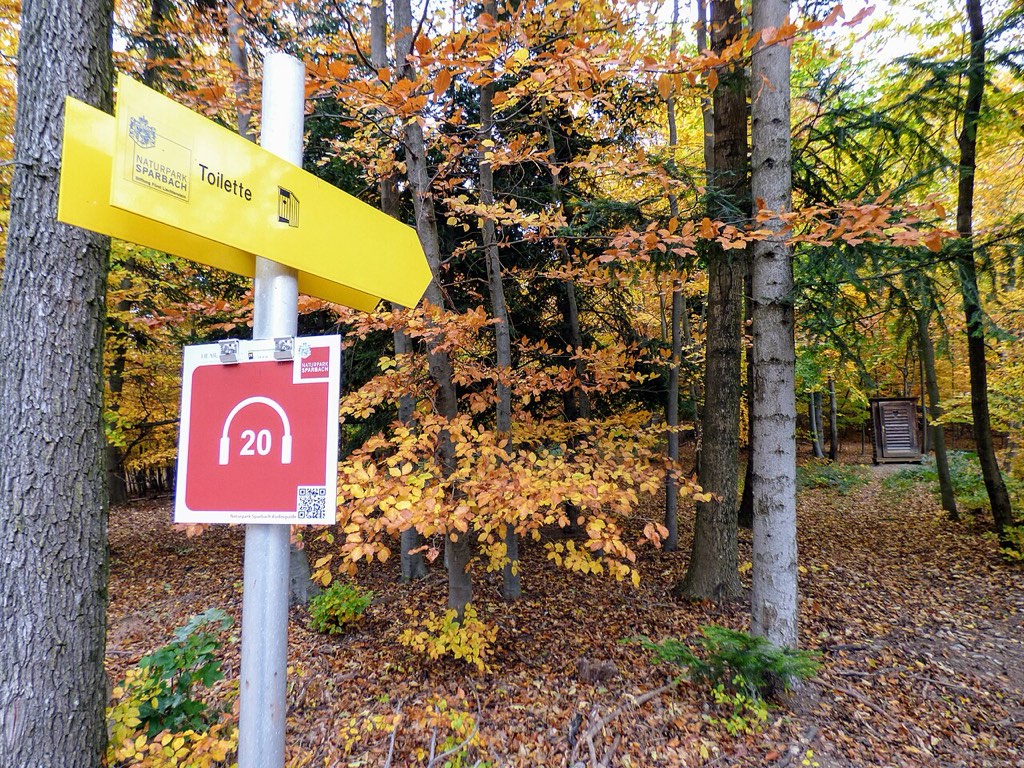 Lesena tabla kaže pot do straniščne hišice v Naravnem parku Sparbach in ima logotip avdiovodnika.